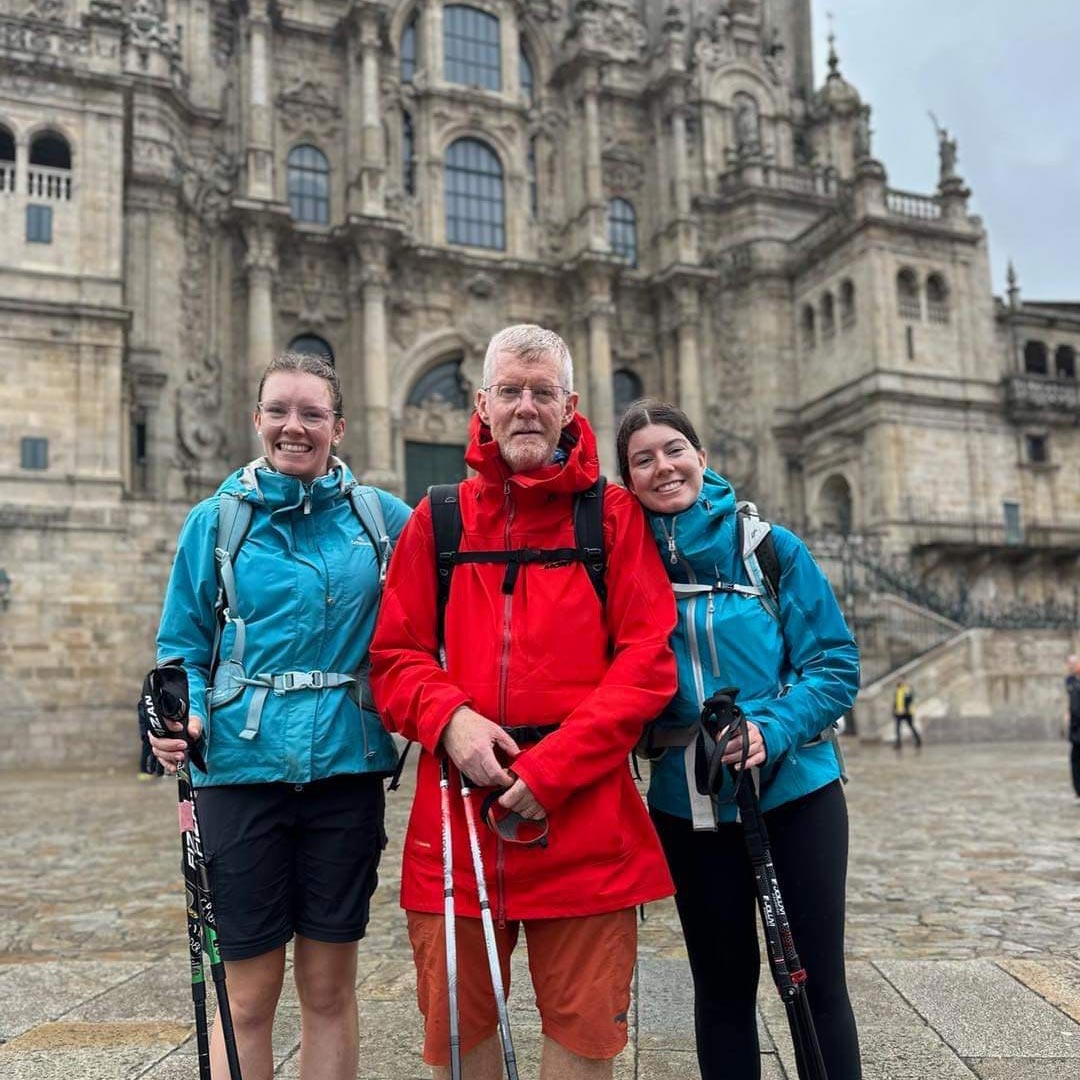 Jeff abroad with his adult daughters. He is in the middle wearing red, they are on either side in blue tops and black pants. All are holding hiking sticks and are posing happily in front of old, European architecture.