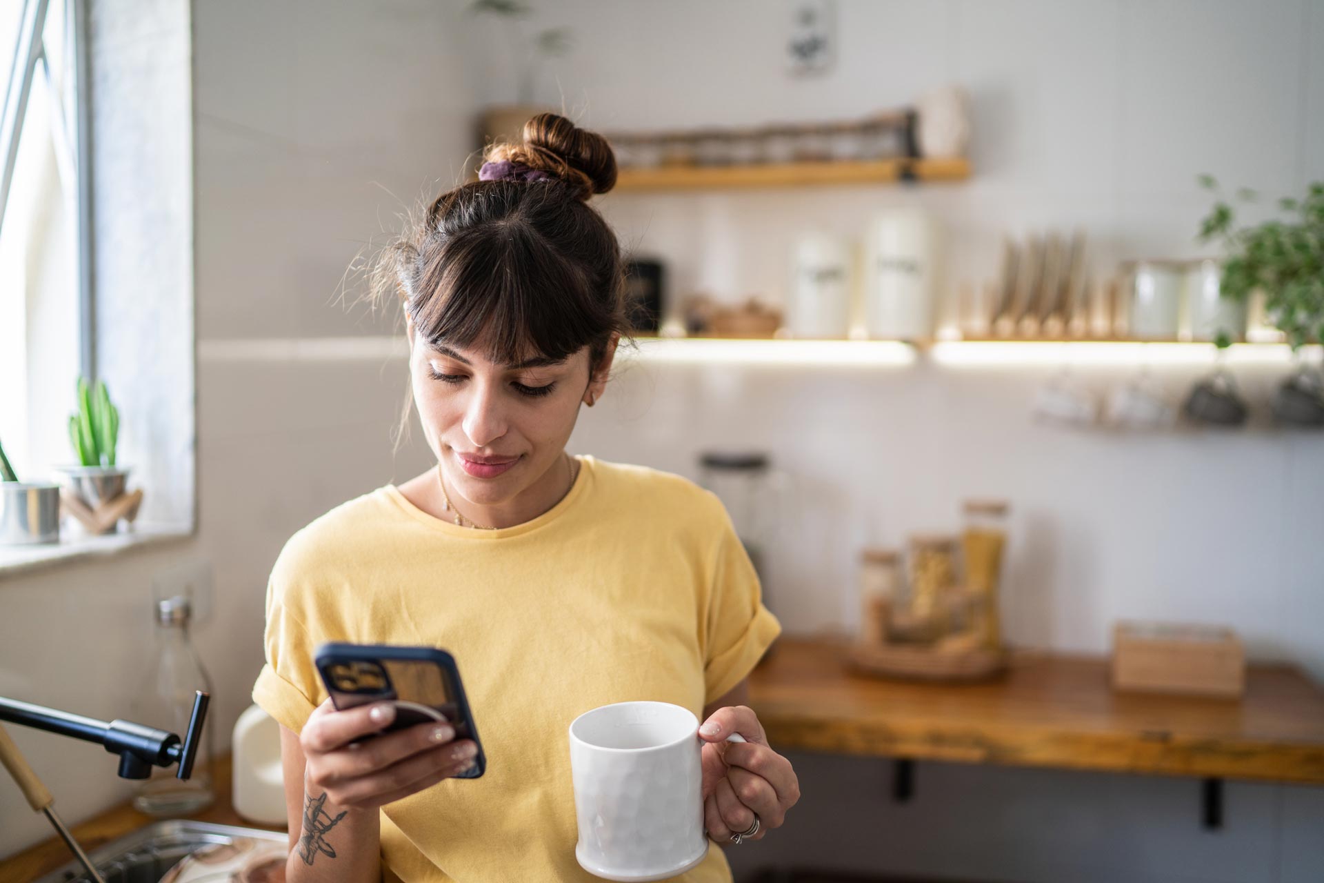 Young woman in yellow enjoying a hot drink and looking at her phone.