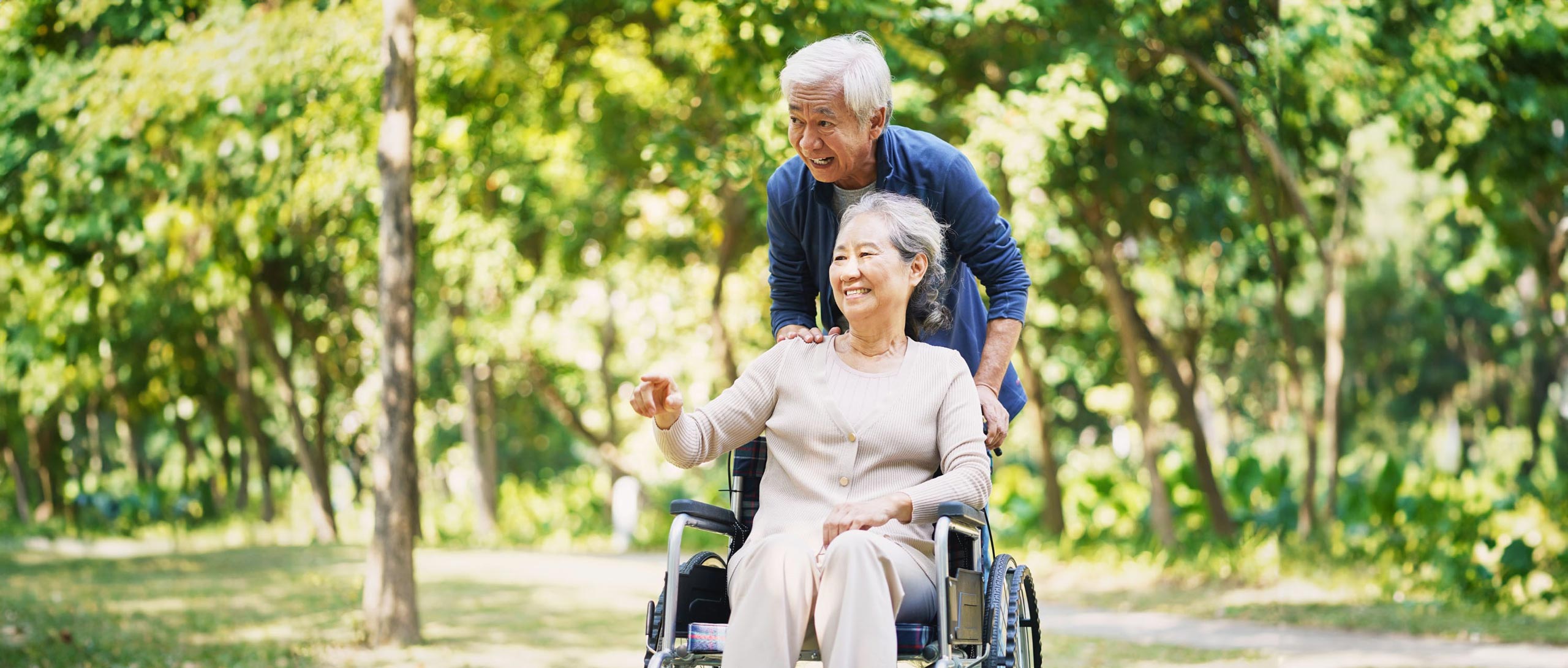 Older couple outdoors, man pushing woman in a wheelchair.